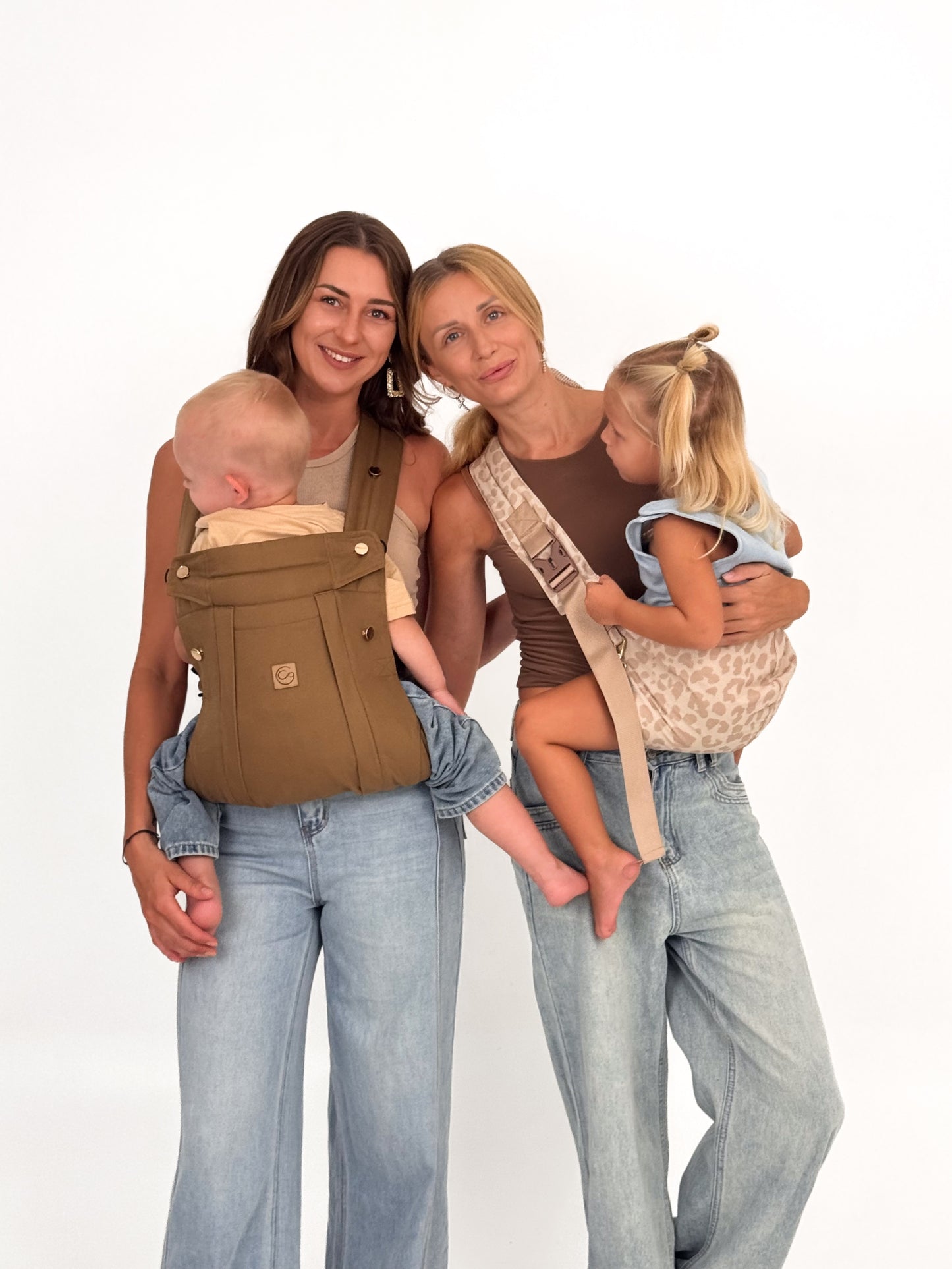 Two mothers standing together, one front-carrying a baby in a Comfort Fit Ergonomic Baby Carrier and the other carrying a toddler in a leopard ring sling, white background