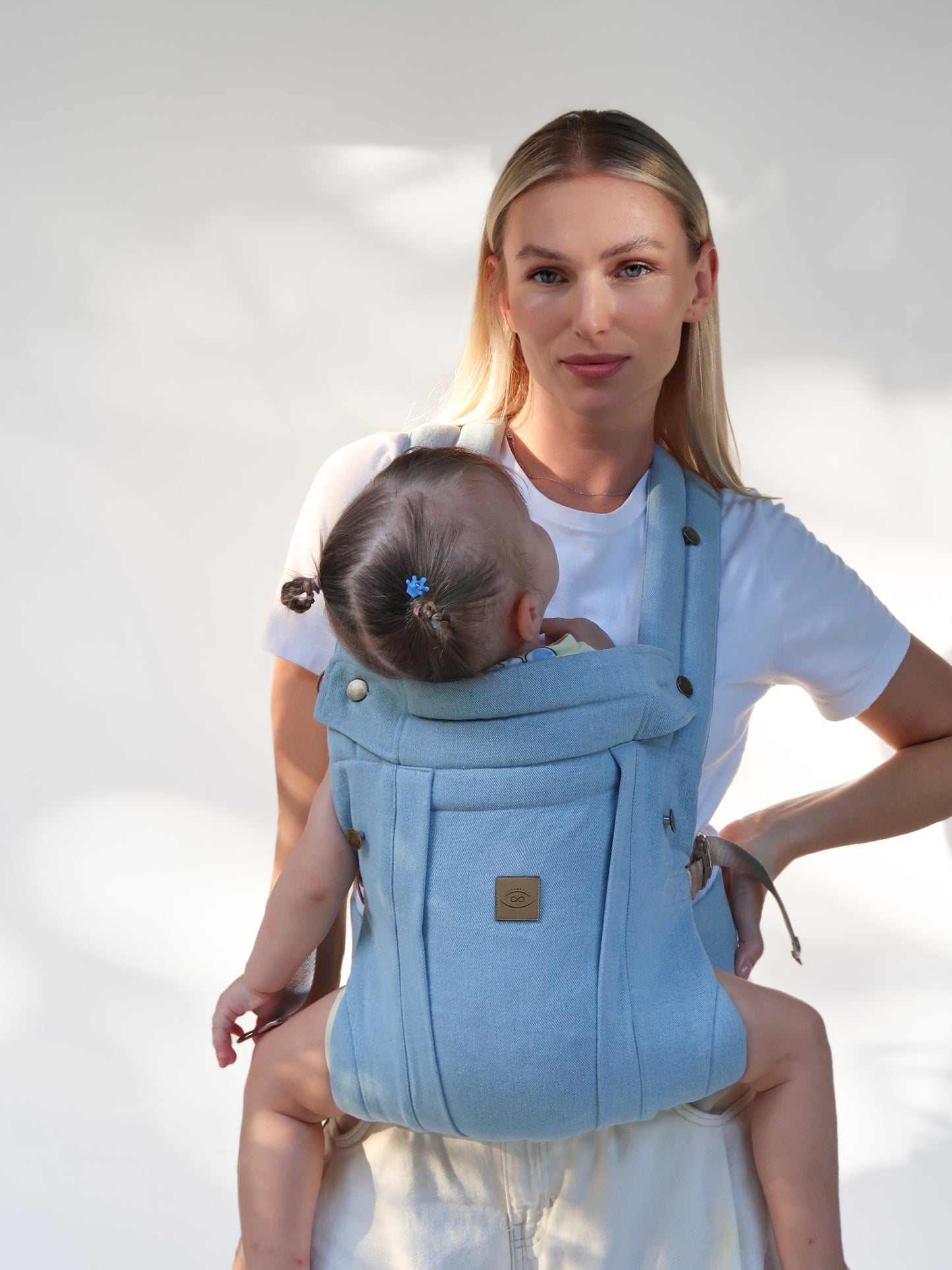 Mother carrying baby in front-carry position in Blue Denim Comfort Fit Ergonomic Baby Carrier, light blue denim carrier with gold logo patch, white background