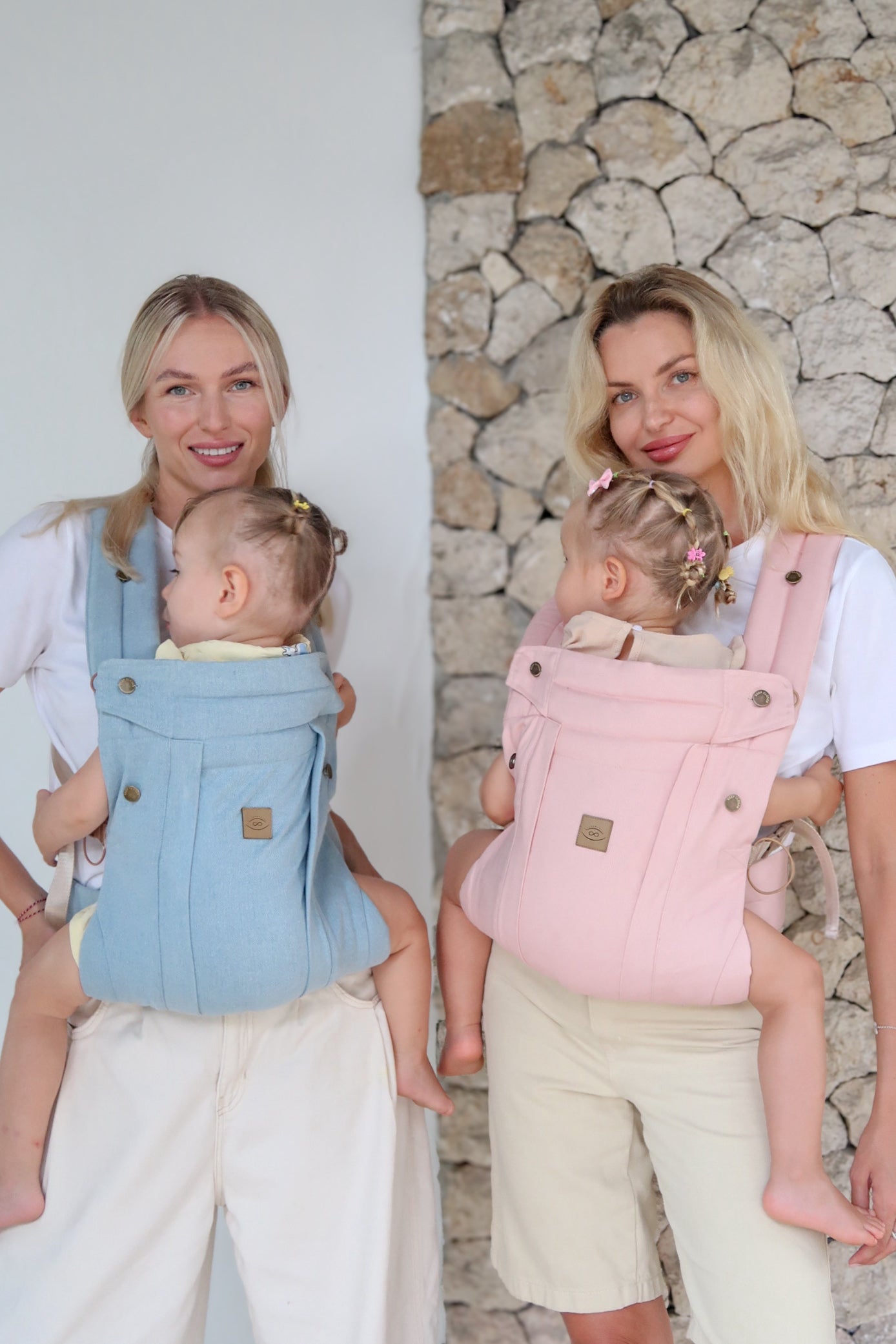 Two mothers carrying babies in Comfort Fit Ergonomic Baby Carriers in blue and pink, front-carry position, stone wall background