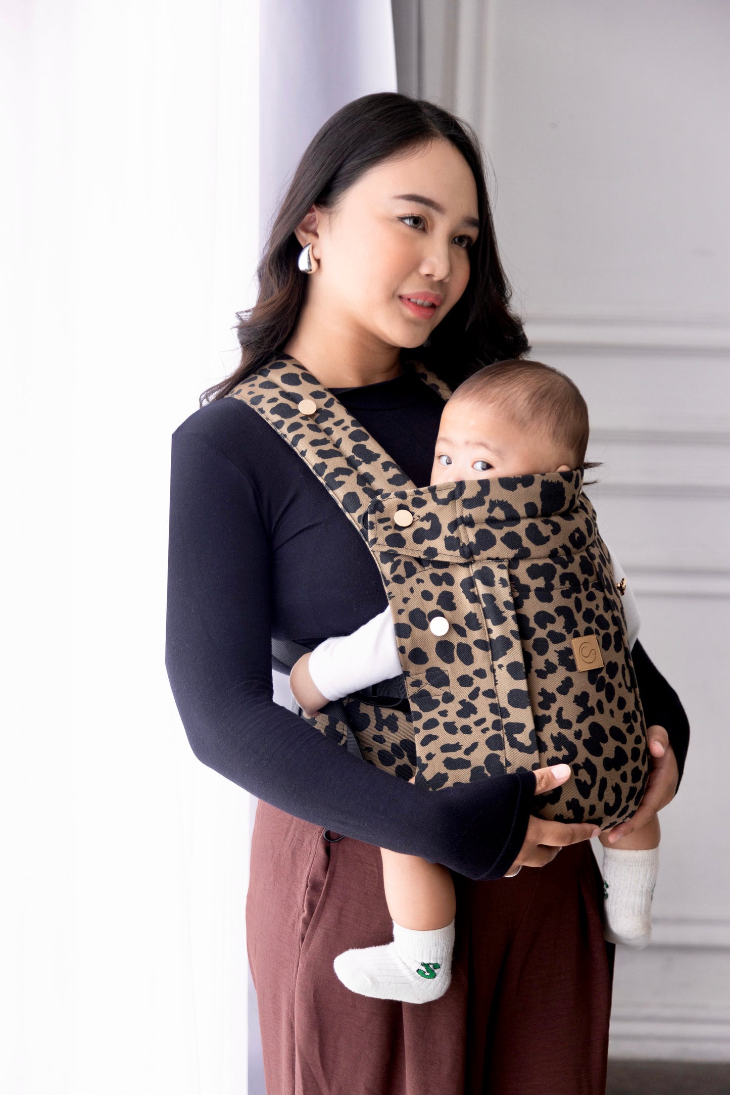 Caramel Leopard Comfort Fit Ergonomic Baby Carrier lifestyle photo, mother carrying baby facing outward in front-carry position wearing caramel leopard print carrier with gold logo patch, white studio background