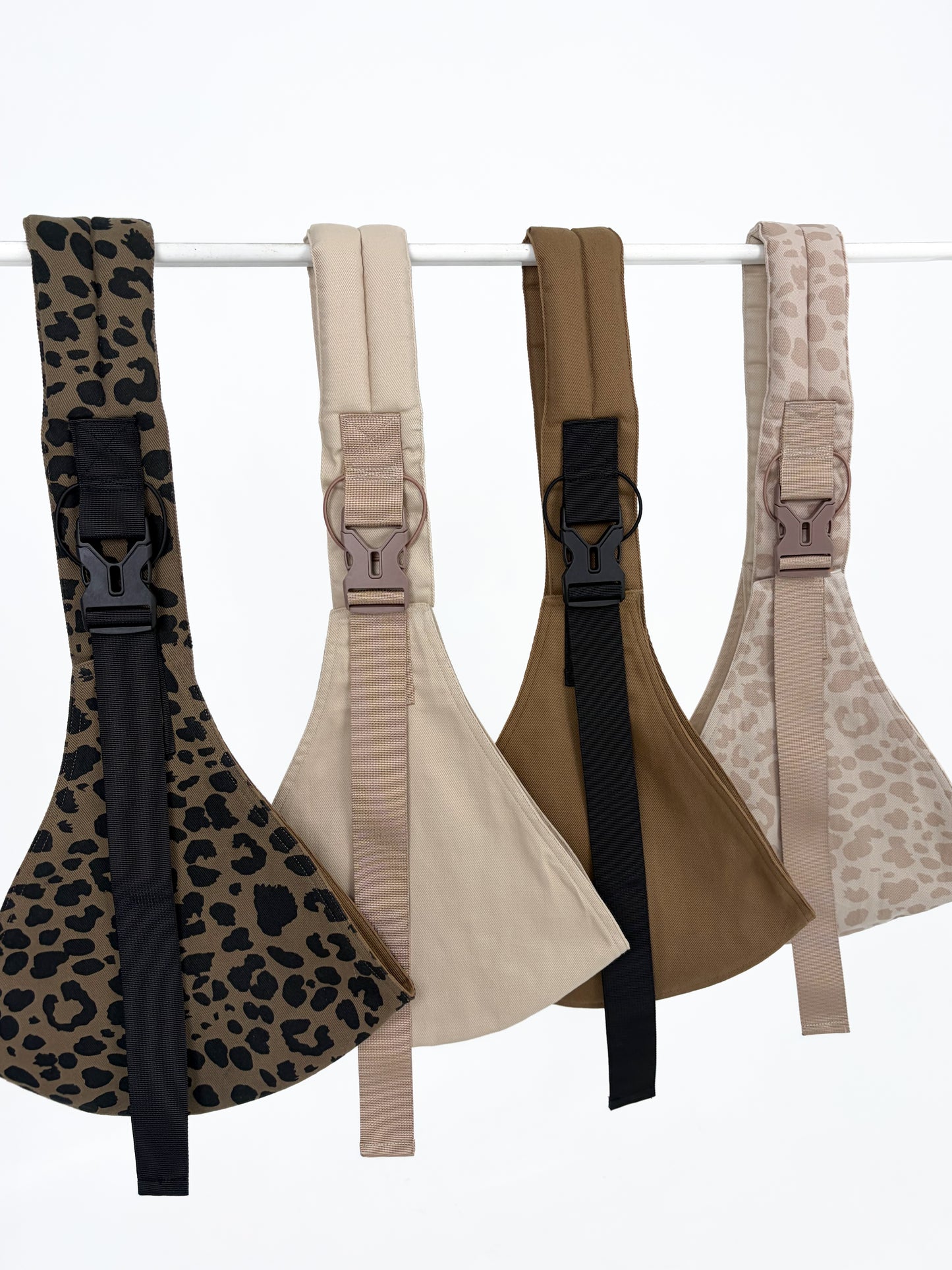 Keep Closer Toddler Hip Carriers in multiple colour variants hanging on a rack, showing leopard print and solid fabric options with black chest straps, white background