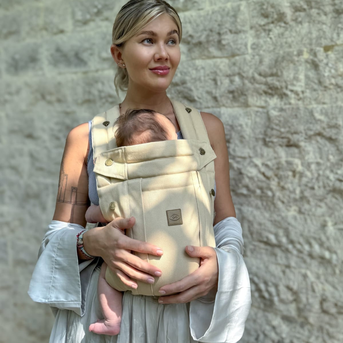 A baby carrier in a sand color, with an infant inside, being worn by a person with a light-colored dress and a waist belt.