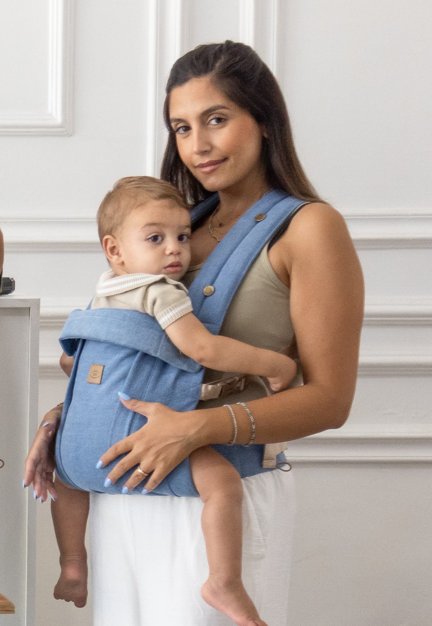 Woman holding a baby in a blue denim carrier against a white wall.