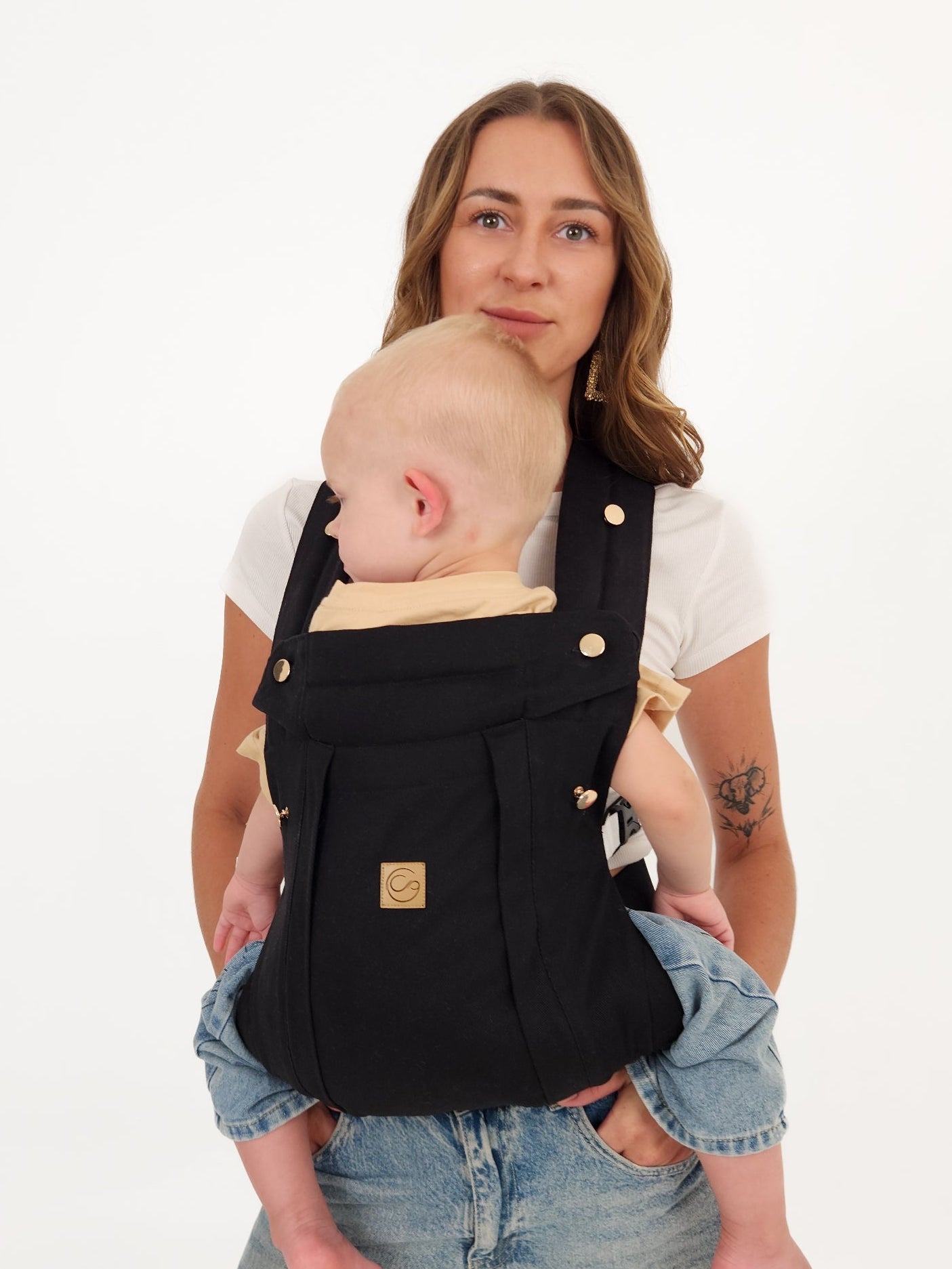 Mother carrying baby in front-carry position in Black Comfort Fit Ergonomic Baby Carrier, black fabric with gold buttons and logo patch, white background