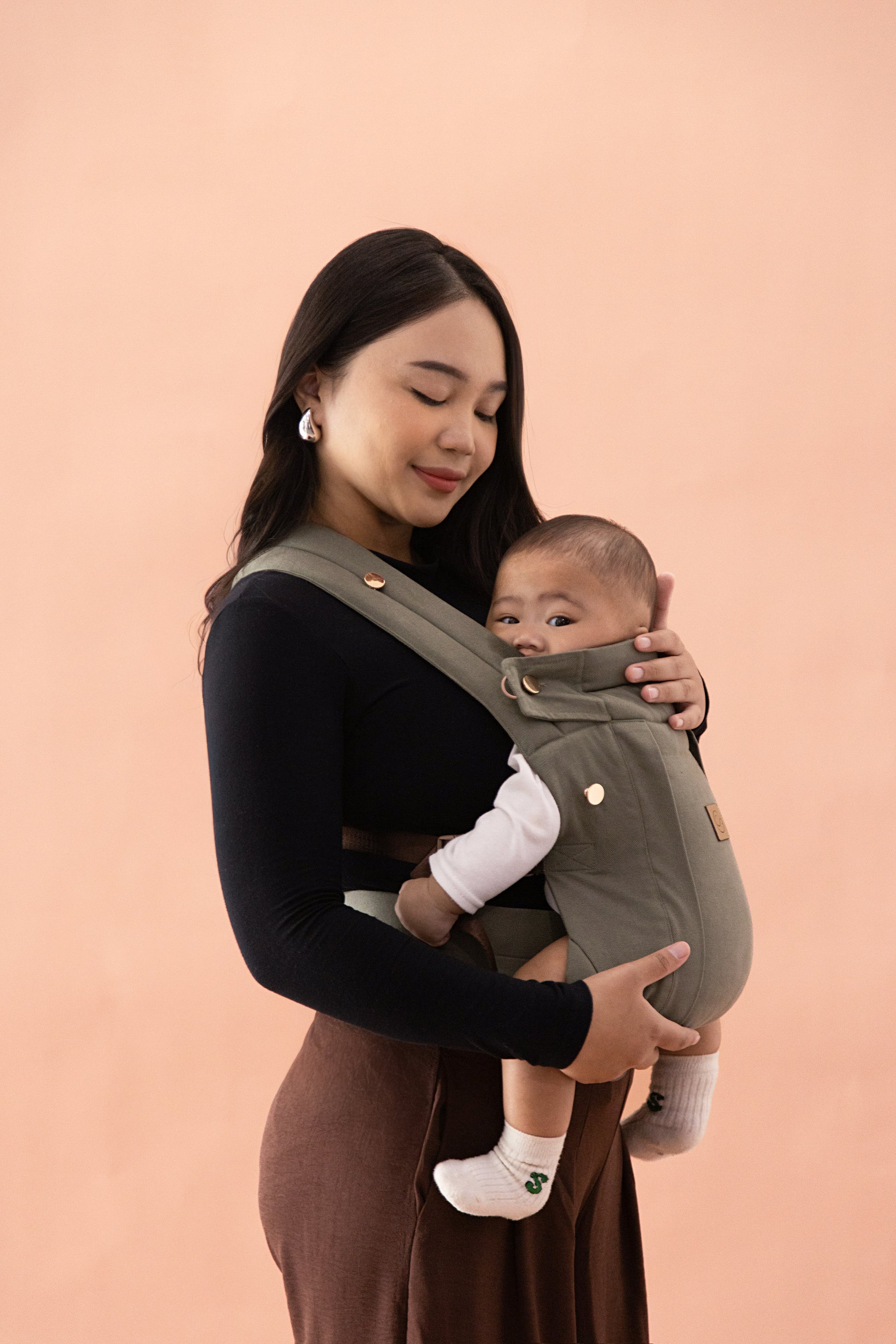 Comfort Fit Ergonomic Baby Carrier lifestyle photo, mother looking down at newborn in front-carry position with detachable hood over baby, olive khaki carrier, pink studio background