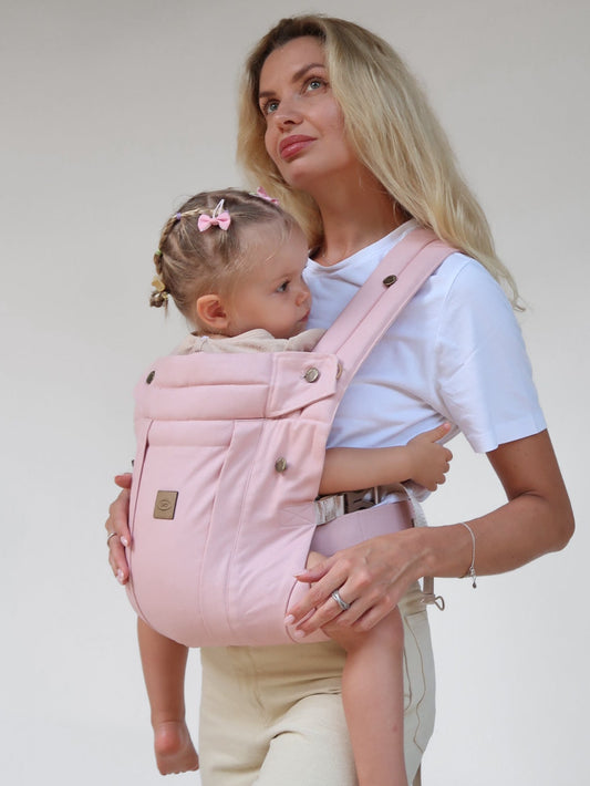 Woman holding a baby in a pink carrier against a plain background