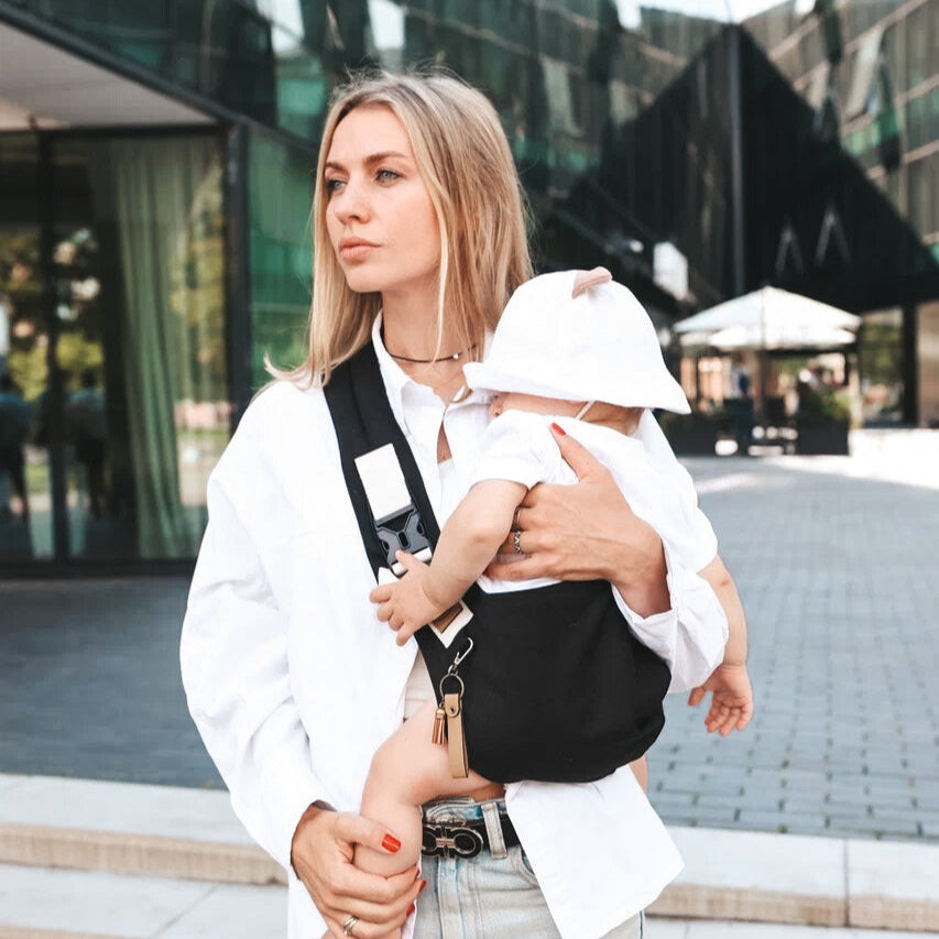 A toddler in a black organic cotton sling carrier with a white hat being carried by a adult outdoors.