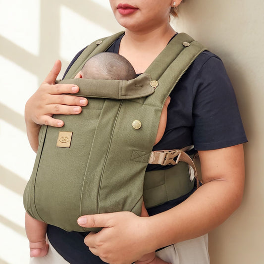 A person wearing a beige baby carrier with a sleeping infant inside.