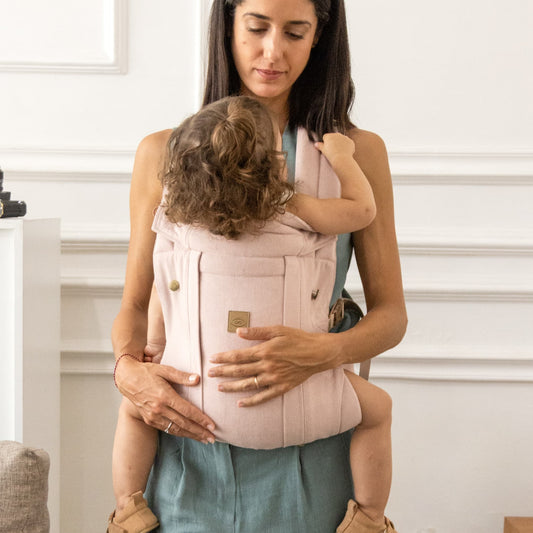 Woman holding a child in a pink baby carrier against a white wall.