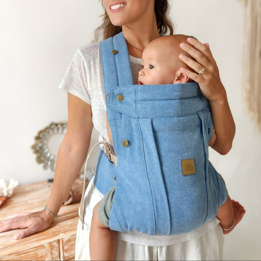 Woman holding a baby in a denim blue baby carrier against a white wall.